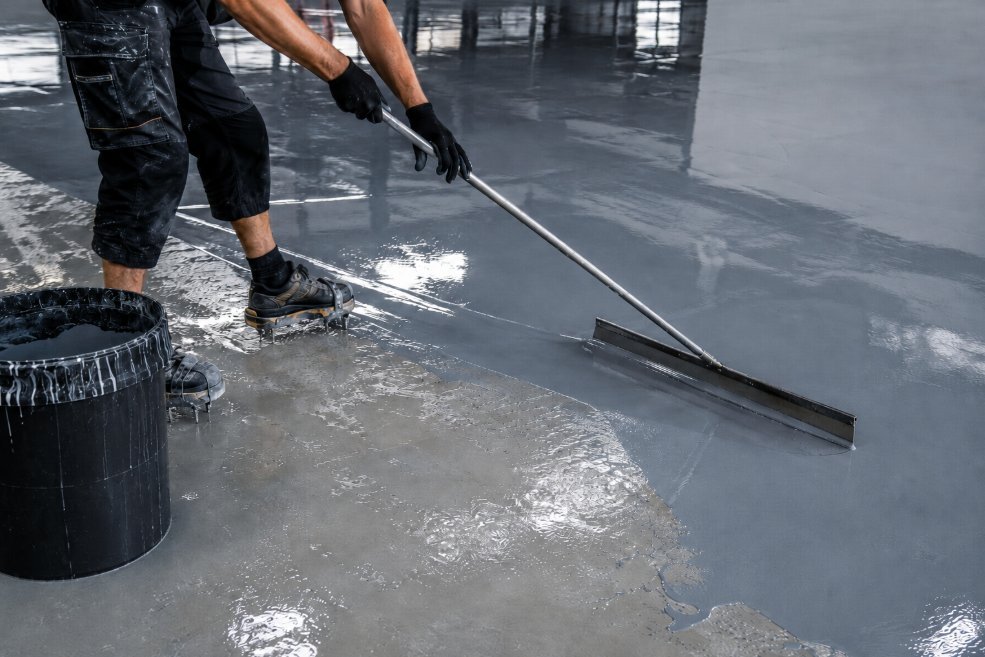 Operario aplicando resina epoxi autonivelante con rastra metálica sobre el suelo, producto líquido extendiéndose en superficie brillante