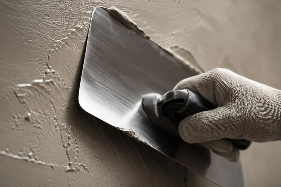 Mano enguantada aplicando microcemento color gris con llana de acero inoxidable sobre pared, técnica de movimiento curvo característico