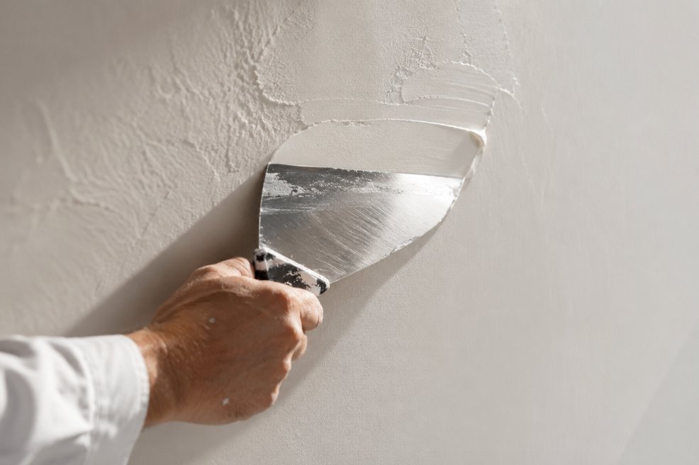 Detalle de mano con espátula metálica ancha aplicando masilla blanca sobre una pared
