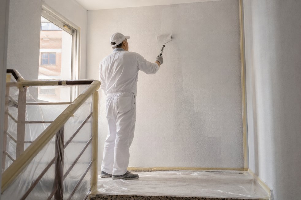 Pintor profesional con mono blanco aplicando pintura con rodillo en la pared de un descansillo de escalera comunitaria con plástico protegiendo el suelo