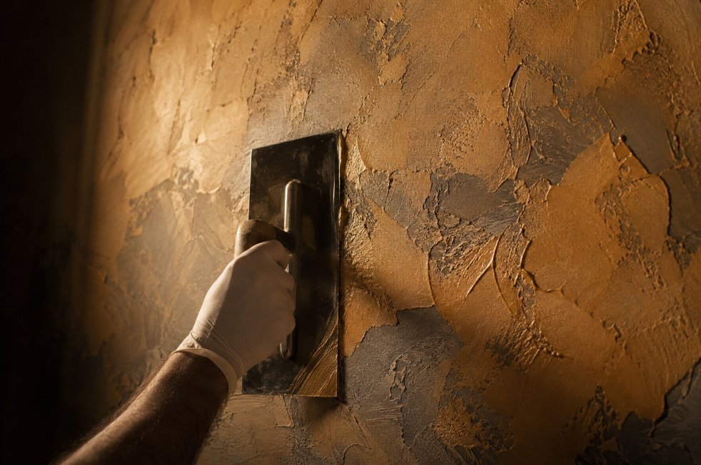 Mano con guante aplicando estuco veneciano con llana sobre una pared, textura ocre dorado con sombras dramáticas