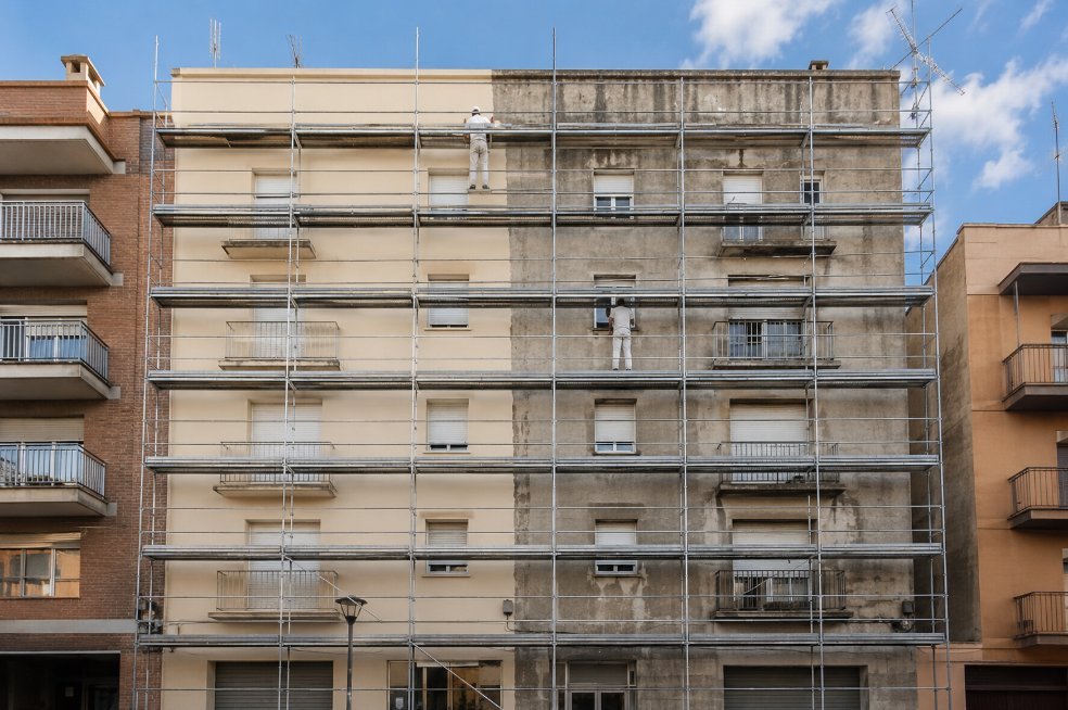 Edificio residencial de cinco plantas en Lleida con andamio europeo montado y la fachada a medio pintar, mitad crema nueva y mitad gris envejecido